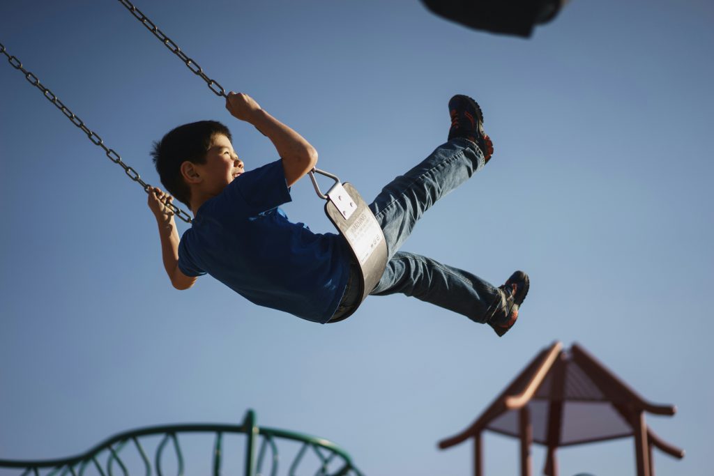 Little boy with black hair in blue shirt and blue jeans swinging on a swing set with the blue sky in the background
